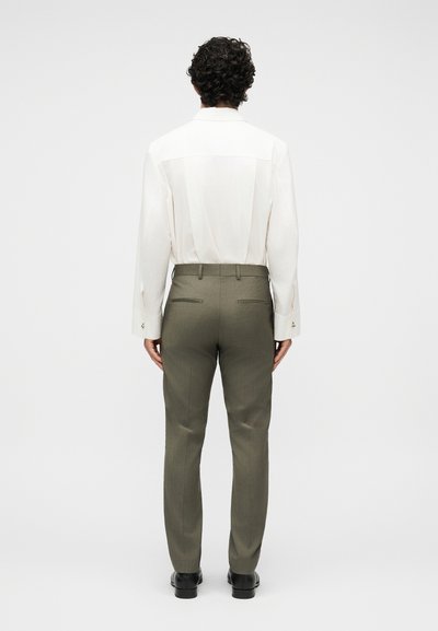 Άνδρας με σγουρά μαλλιά, που φοράει λευκό πουκάμισο με μακριά μανίκια και παντελόνι φόρμας σε ελαιόχρωμο πράσινο, στέκεται γυρισμένος με την πλάτη σε απλό φόντο.