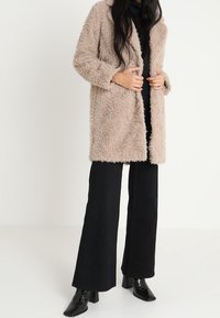 Miękki beżowy płaszcz o luźnym kroju, z długimi rękawami i bocznymi kieszeniami, zestawiony z czarnymi spodniami o szerokich nogawkach oraz błyszczącymi czarnymi botkami do kostek.