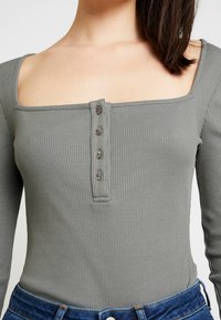 Grå rippad långärmad topp med fyrkantig halsringning, prydd med en knappslå vid bröstet och en figurnära passform.