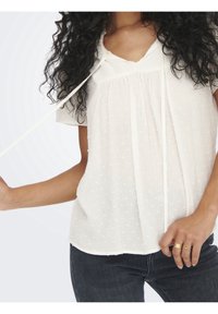 Valkoinen pusero, jossa lyhyet hihat, kohokuvioitu kangas, jossa pieniä koholla olevia pilkkuja, V-pääntie ja solmittava yksityiskohta kauluksessa.