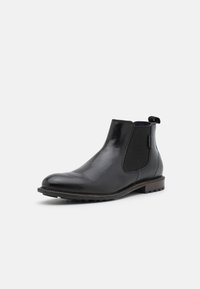 Botín Chelsea de cuero negro con paneles laterales elásticos, punta redondeada y suela de goma. Incluye un tirador para facilitar su uso. Textura suave.