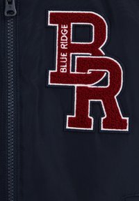 Marinblå jacka med stora röda och vita broderade bokstäver "BR" och vertikalt text "BLUE RIDGE", bredvid en svart dragkedja.