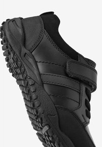 Chaussures de sport noires avec une sangle en velcro, dotées d'une semelle en caoutchouc texturé, d'un dessus en synthétique lisse et d'un col rembourré pour le confort.
