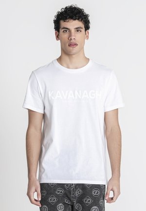 Junger Mann mit lockigem Haar, der ein weißes "KAVANAGH"-T-Shirt und dunkle gemusterte Hose trägt, steht vor einem schlichten hellen Hintergrund.