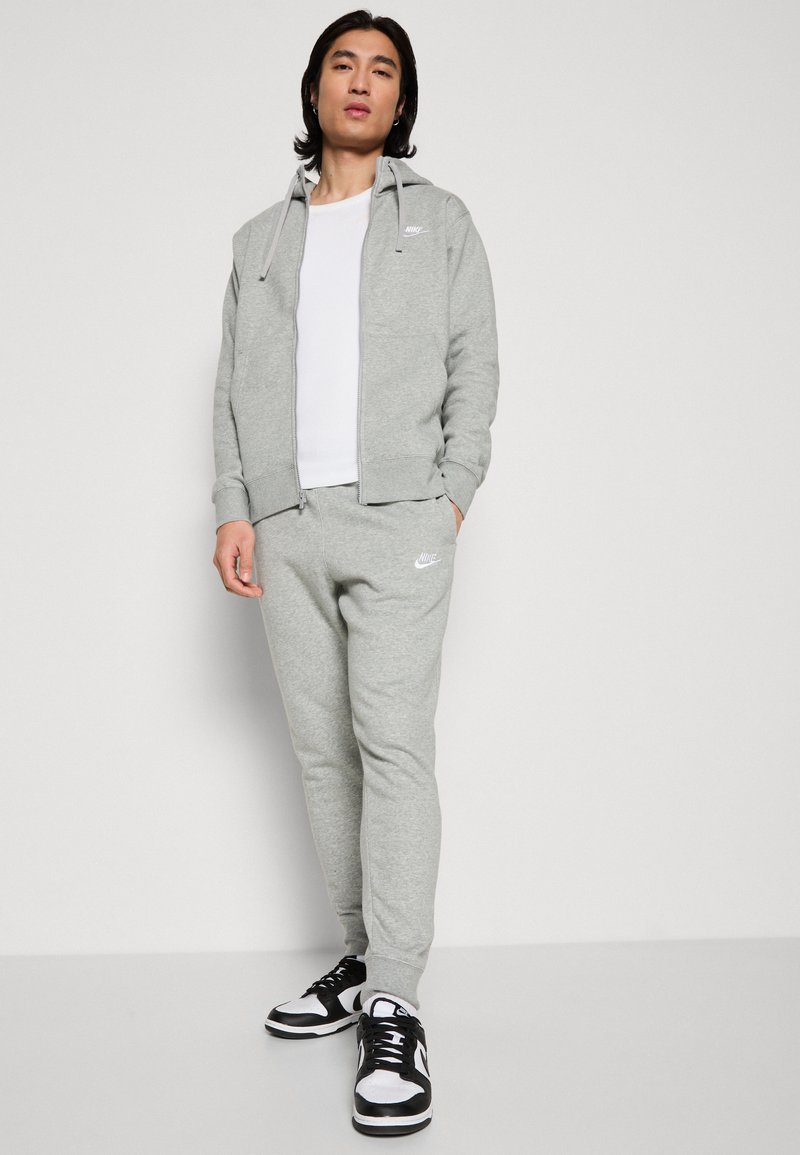 Sweat à capuche gris Nike avec fermeture éclair et logo, associé à un pantalon de jogging assorti. Des baskets noir et blanc complètent l'ensemble. Texture lisse.