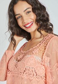 Top de croché en melocotón que presenta patrones intrincados, tejido transparente y un escote redondo, con mangas largas y acentos florales.