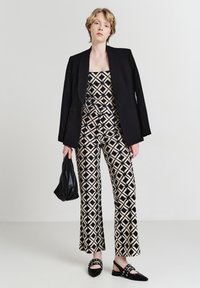 Une personne se tient debout portant un blazer noir sur une combinaison à motifs géométriques beige et noire, tenant un sac noir et portant des chaussures à pointes noires.