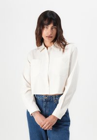 Camisa blanca recortada de botón en una tela suave, con dos bolsillos frontales y un detalle de corazón en relieve en el bolsillo izquierdo.