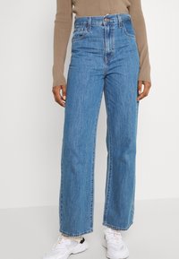 Person som bär högmidjade, raka blå jeans, en beige långärmad topp och vita klumpiga sneakers, stående mot en vit bakgrund.