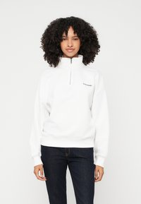 Donna con capelli ricci che indossa una felpa bianca con zip a metà e il logo "Guess Jeans" e jeans blu scuro, in piedi davanti a uno sfondo semplice.