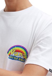 Person, der ein weißes T-Shirt mit einem Regenbogen und dem "Vans off the wall"-Logo auf der Brust trägt, mit verschränkten Armen.