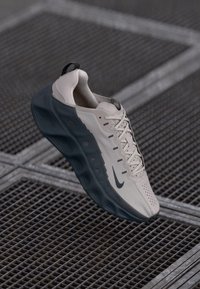 Beige sporta apavi ar teksturētu virsu, tumšu zoli un unikālu viļņveida dizainu. Iekļauj auklas un mazu logo sānos.