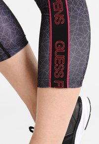 Svarta och grå träningstights med geometriskt mönster; har en sidopanel med röd "GUESS FIT" text, elastisk midja och sydda detaljer.