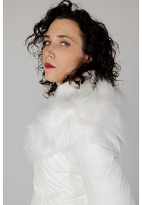 Veste doudoune blanche avec une capuche en fausse fourrure, texture matelassée et détail de logo sur la manche. Design élégant avec un col arrondi.