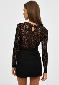 Top de encaje negro con mangas largas, patrón floral y detalle en la parte posterior con abertura, combinado con una falda negra ajustada. Apariencia texturada y semi-translúcida.