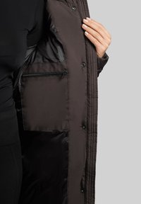 Pruun isoleeritud jakk, millel on tikandite tekstuur, näidates sisetaskut, millel on lukk ja nööpklambrid ees.