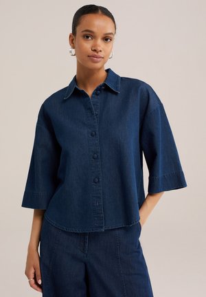 Femme portant une chemise en jean bleu foncé à boutons avec manches trois-quarts et un pantalon en jean taille haute assorti, debout avec la main dans la poche.