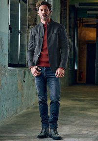 Gray button-up jacket over a red ribbed sweater, paired with blue jeans and black boots. The setting features textured walls and natural light.