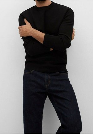 Homme portant un pull à manches longues noir et un jean bleu foncé, debout les bras croisés devant un fond clair uni.