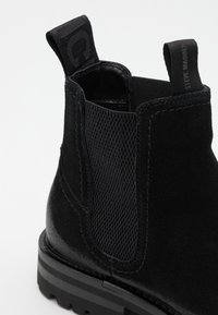 Bottes Chelsea en daim noir avec panneau latéral élastique, languettes côtelées et semelle épaisse à rainures, mettant l'accent sur la cheville et le talon.