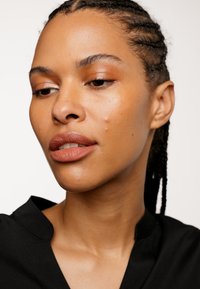 Närbild av en person med flätat hår, iklädd en svart topp. Drag som inkluderar varma toner i sminket, markerade kindben och en jämn hudtextur.