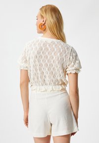 Top blanco de punto y manga corta con patrón texturizado y dobladillo con volantes, combinado con pantalones cortos color crema. La modelo lleva grandes pendientes de aro naranjas.
