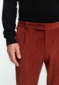 Rostfarbene Cordhosen mit vertikalen Streifen, ausgestattet mit einem Knopfverschluss und einer gefalteten Vorderseite. Kombiniert mit einem schwarzen Pullover.