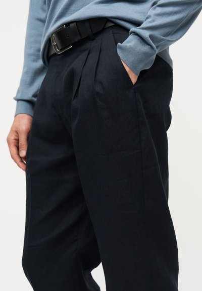 Άνδρας που φοράει μαύρο πιετέ παντελόνι με πλαϊνές τσέπες, μαύρη ζώνη και μπλε πουλόβερ με μακριά μανίκια, με το χέρι στην τσέπη.