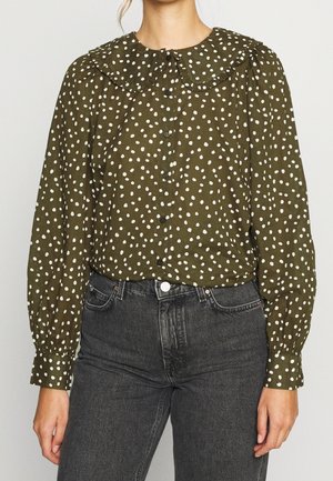 Olijfgroene blouse met witte onregelmatige polkadots, wijde kraag met ruches, knoopsluiting aan de voorkant en lange pofmouwen, gecombineerd met zwarte jeans met hoge taille.