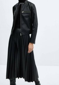 Veste en cuir noir avec un col haut et des zip métalliques, assortie à une jupe plissée noire. Le tout est conçu dans une silhouette épurée.