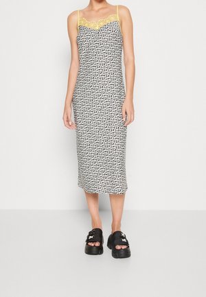 Femme portant une robe midi fleurie noire et blanche avec un bord en dentelle jaune et des bretelles fines, associée à des sandales compensées noires.