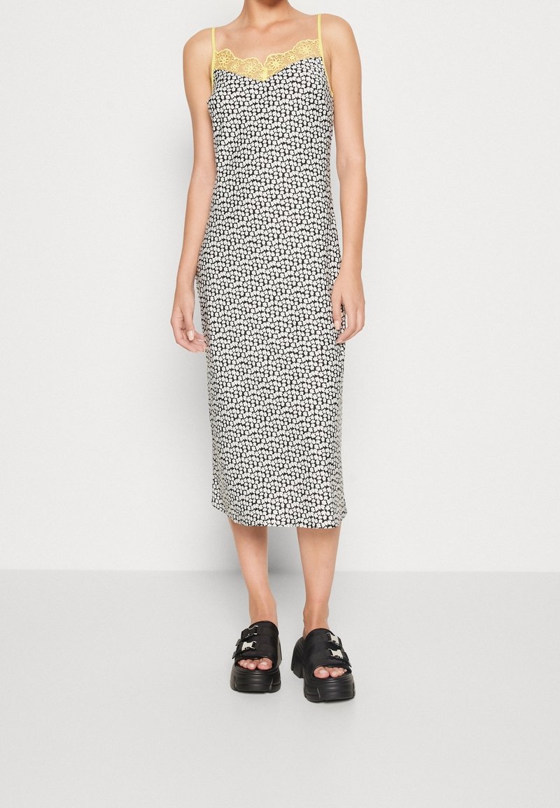 Femme portant une robe midi fleurie noire et blanche avec un bord en dentelle jaune et des bretelles fines, associée à des sandales compensées noires.