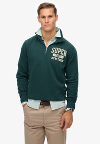 Felpa zipata verde scuro con la scritta "SUPER DRY NEW YORK" stampata in crema, indossata sopra una camicia a righe blu chiaro, abbinata a pantaloni di un leggero beige.