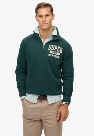 Dunkelgrüner Zip-Hoodie mit "SUPER DRY NEW YORK" in creme bedruckt, übereinander getragen mit einem hellblau gestreiften Hemd, kombiniert mit hellbraunen Hosen.