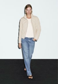 Beige gewatteerde jas met een hoge kraag, gedragen over een lichte top, gecombineerd met losse blauwe jeans en zwarte sandalen op een donkere achtergrond.