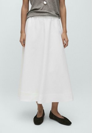 Kvinde iført grå ærmeløs top, hvid ankel-lang nederdel, sorte ballet-sko og vedhængs-halskæde mod en ensfarvet baggrund.