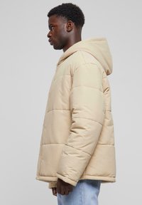 Beige pufferjacka med huva, som har kviltade paneler och dragkedjestängning. Tyget har en mjuk textur, lämplig för kallt väder.