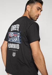 Schwarzes Baumwoll-T-Shirt mit einem grafischen Design, das den Text "FOREVER FORWARD" und einen Fußball zeigt, akzentuiert mit blauen Sternen und Streifen.