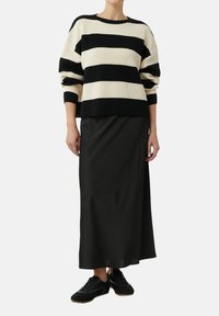 Woman wearing a black and white wide striped sweater, black long skirt, and black sneakers standing against a white background.