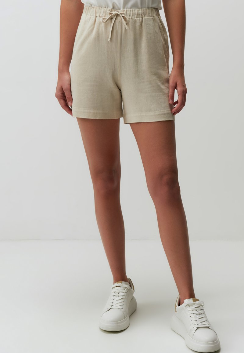 Beige shorts gemaakt van een lichtgewicht stof met een elastische tailleband en drawstring. Gecombineerd met witte sneakers op een neutrale achtergrond.