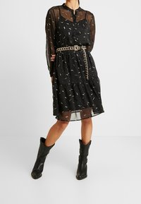 Svart genomskinlig klänning med gyllene prickar, lagerdesign, långa ärmar och ett bälte. Matchas med knähöga svarta läderstövlar.