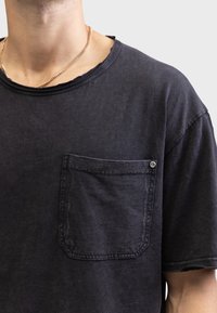 Sort bomulds T-shirt med rund halsudskæring, rå kanter og en brystlomme med trykknapdetalje. Blødt, struktureret stof.