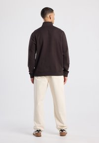 Dark brown zip-up sweatshirt with a high collar, paired with light beige pants. Features long sleeves and ribbed cuffs. Skin-tone sneakers complete the look.