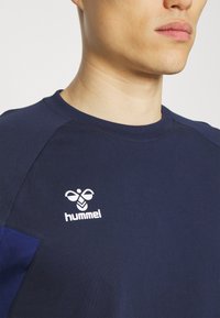 Mörkblå t-shirt med runda halsringningar, tillverkad av mjukt material, med en kontrasterande blå panel och vit "hummel"-logga på bröstet.