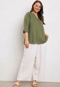 Donna sorridente con lunghi capelli castani, che indossa una camicetta verde oliva con bottoni, pantaloni bianchi larghi e sandali a piattaforma bianchi, in piedi contro un muro bianco.