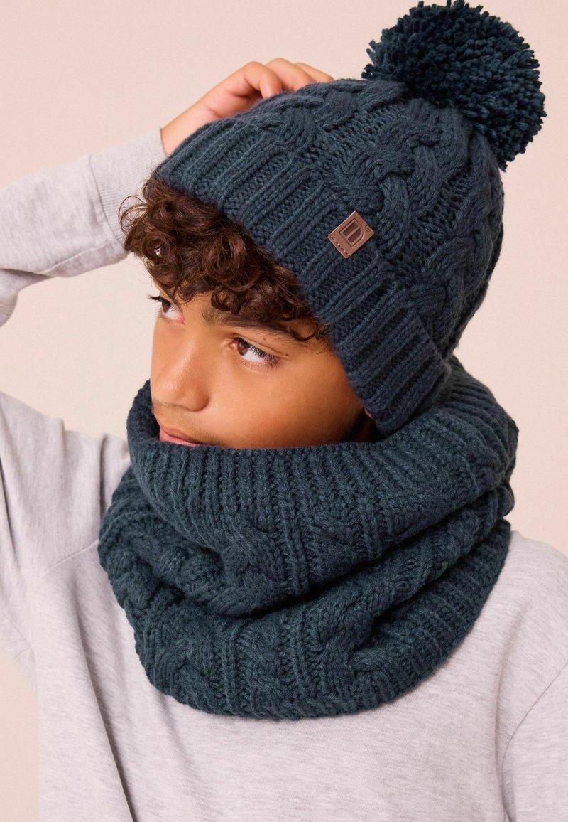 Bonnet tricoté en vert foncé avec un pompon et un design en câble texturé ; écharpe cowl assortie en tissu épais à maille côtelée.