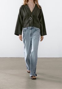 Giacca corta verde scuro con bottoni e maniche larghe, abbinata a jeans azzurri a gamba dritta e sandali bassi. Accessori ridotti al minimo.