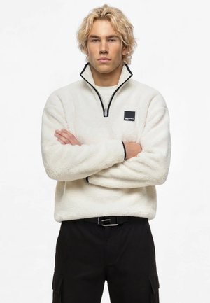 Homme blond, les bras croisés, portant un pull polaire blanc avec une fermeture éclair noire et un pantalon noir sur fond blanc uni.