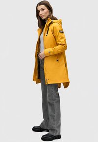 Gele waterdichte regenjas met capucho, drukknoopsluiting en trekkoord. Gecombineerd met grijze jeans en zwarte chunky laarzen.
