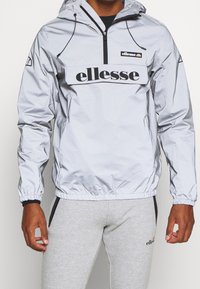 Světle šedý větrový kabát s kapucí a půlzipem v přední části, elastické manžety a výrazné černé logo "ellesse" na hrudi. Hladká, lesklá textura.